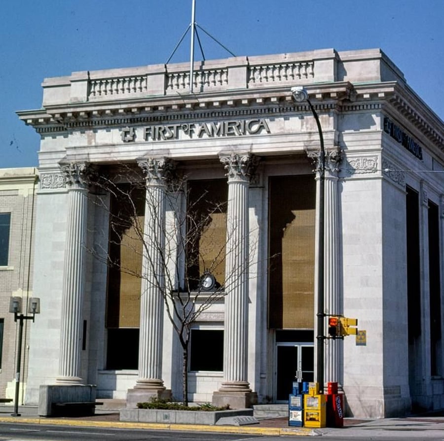 A neoclassical bank building with tall columns and "First America" signage, featuring a leafless tree in front, newspaper boxes on the corner, and traffic lights at the street intersection.