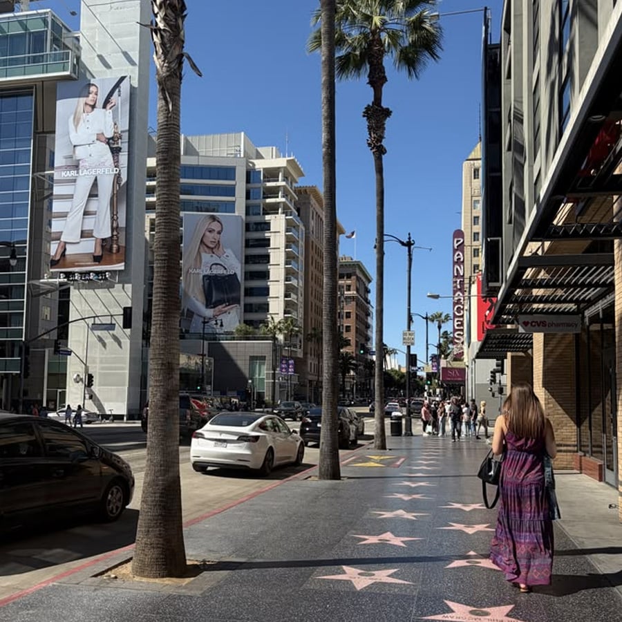 A sunny street scene on the Hollywood Walk of Fame, showing palm trees, star plaques on the sidewalk, cars, pedestrians, and tall buildings with large fashion advertisements in the background.