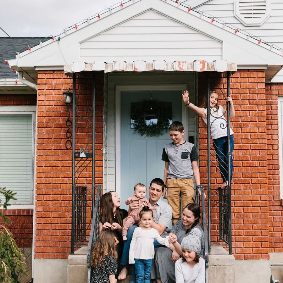 A family of eight poses on the front steps of a brick house. Two children stand on the porch railings, while the others and two adults sit or kneel on the steps, smiling and interacting happily.