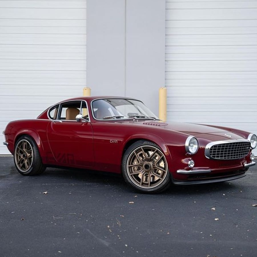 A classic red two-door coupe car with bronze wheels is parked in front of a white industrial garage door on a blacktop surface. The car has a vintage design and shiny exterior.
