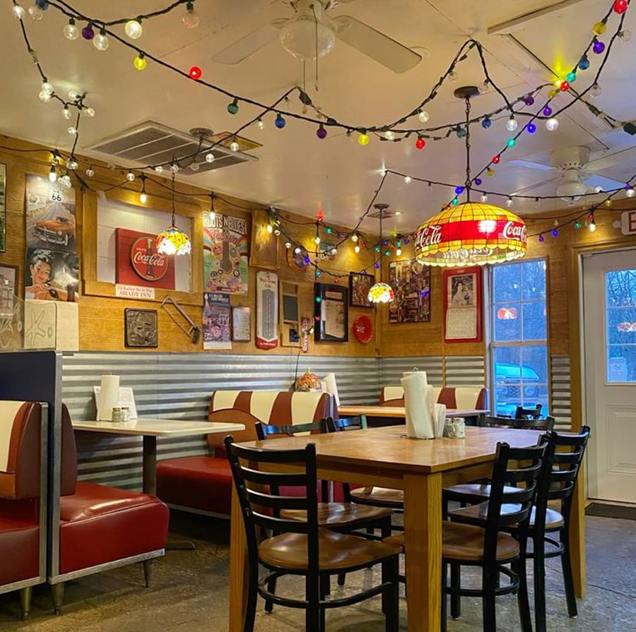 Cozy retro diner with red and white booths, wooden tables, black chairs, and colorful string lights hanging from the ceiling. Walls are decorated with vintage posters and Coca-Cola signs. Natural light comes through a window and door.