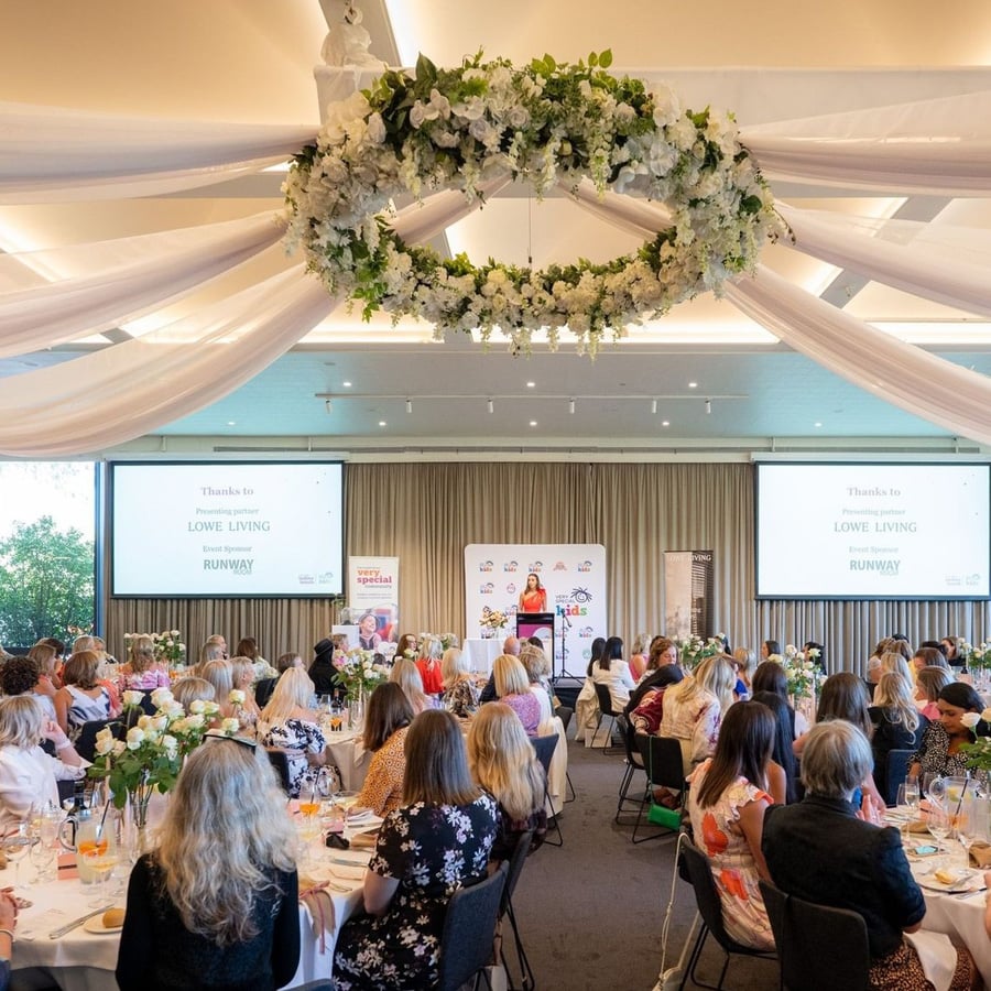 A large group of people sit at decorated round tables during a formal event in a bright banquet hall with floral arrangements, draped white fabric, and a large flower wreath hanging from the ceiling. A speaker stands at a podium.