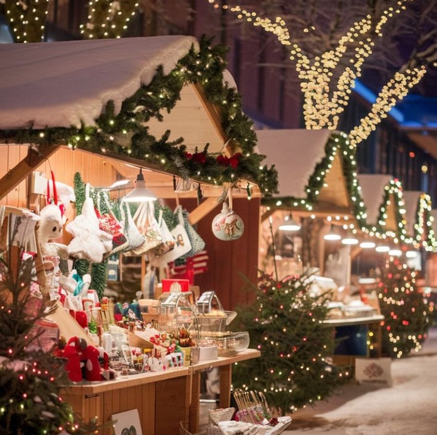 A festive outdoor Christmas market features wooden stalls decorated with garlands and lights, selling holiday gifts and ornaments. Snow covers the ground and rooftops, with trees wrapped in twinkling string lights.