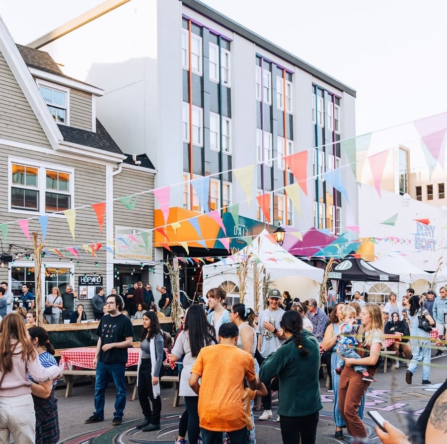 A lively outdoor street festival with people of all ages socializing under colorful bunting flags, surrounded by booths and tall buildings in the background. Some people are dancing and children are playing.