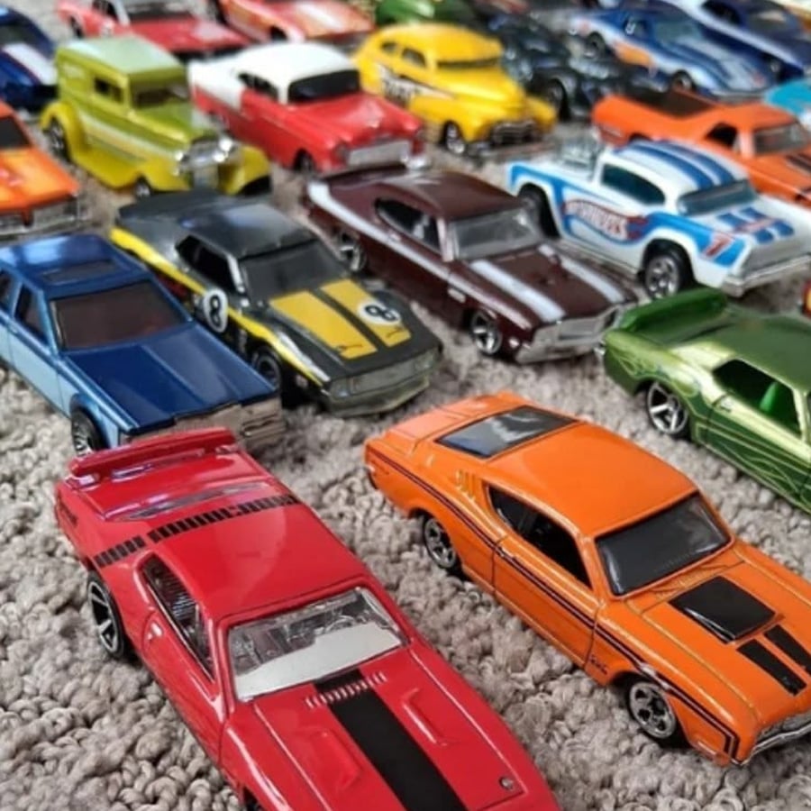 A collection of colorful toy cars, including red, orange, blue, green, and yellow models, is arranged in rows on a textured carpet. The cars have various designs and racing stripes.