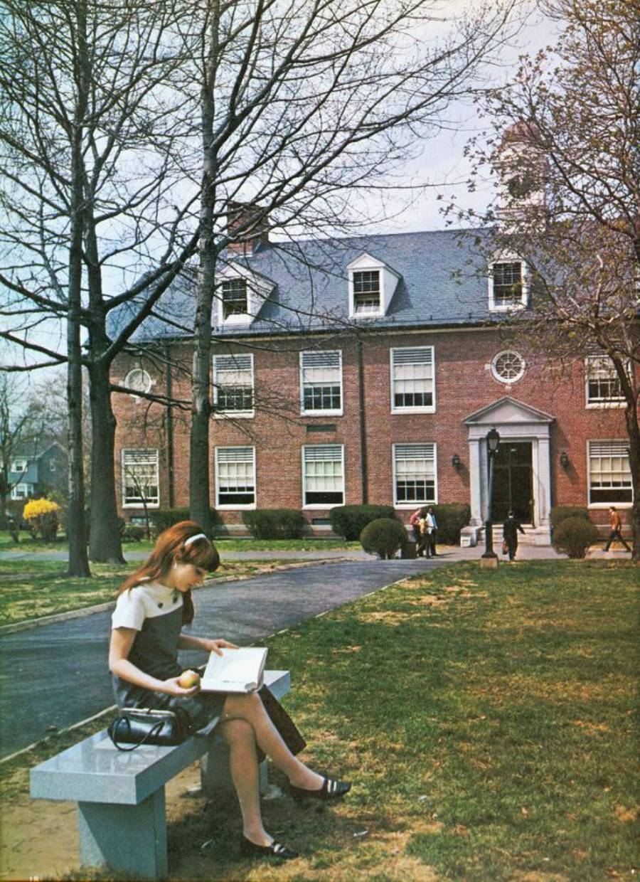 A young woman sits on a bench reading a book and holding an apple on a grassy area in front of a brick academic building with multiple windows and trees, while other people walk nearby.