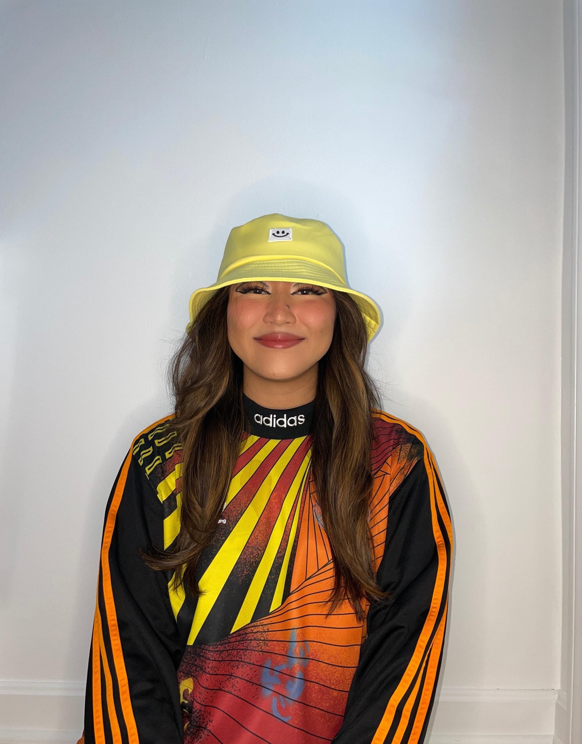 A person with long brown hair wears a yellow bucket hat and a colorful Adidas sports top with orange and yellow stripes, smiling in front of a plain white background.