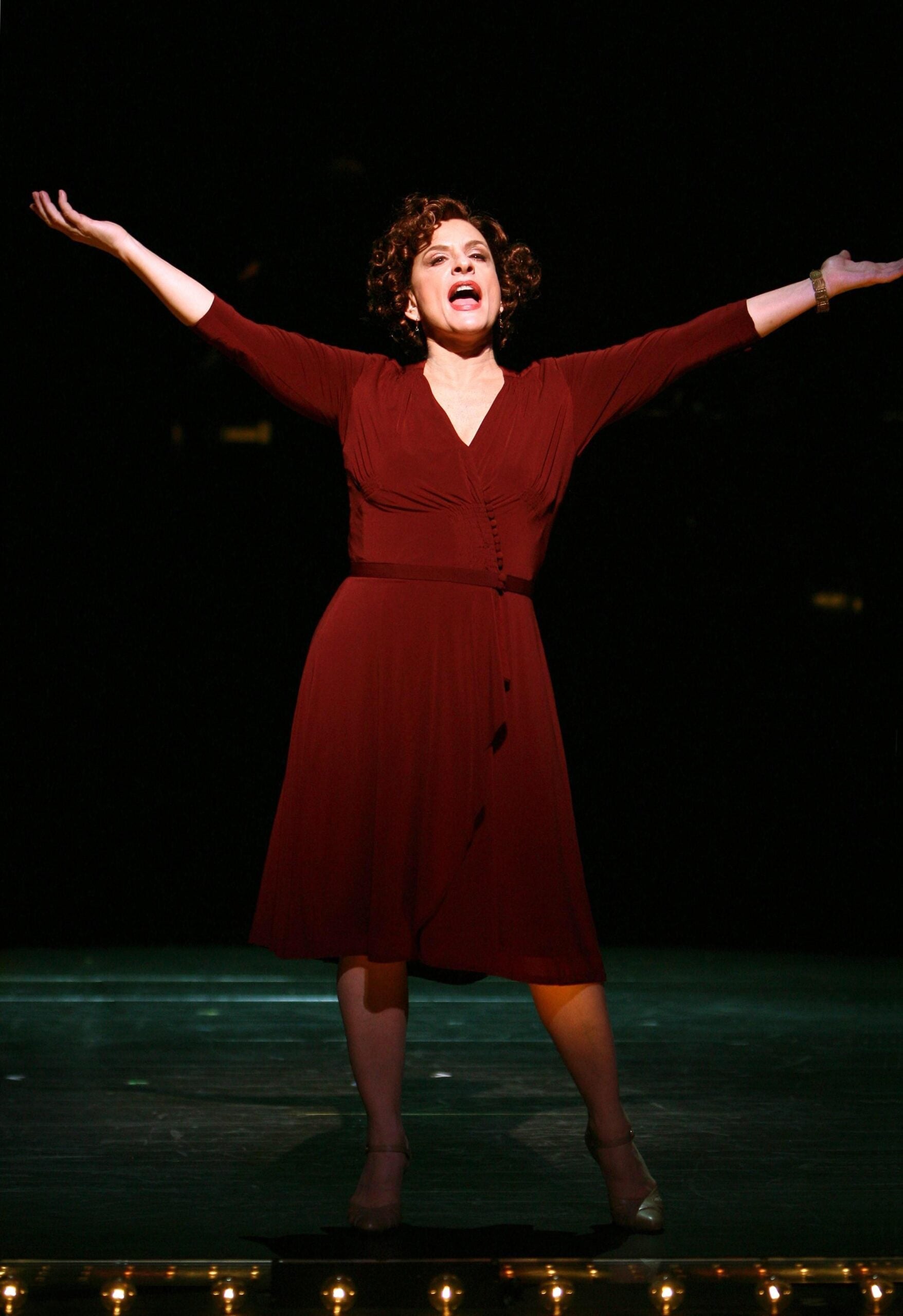 A woman in a red dress stands on stage with arms outstretched, mouth open as if singing, under dramatic lighting with a dark background.