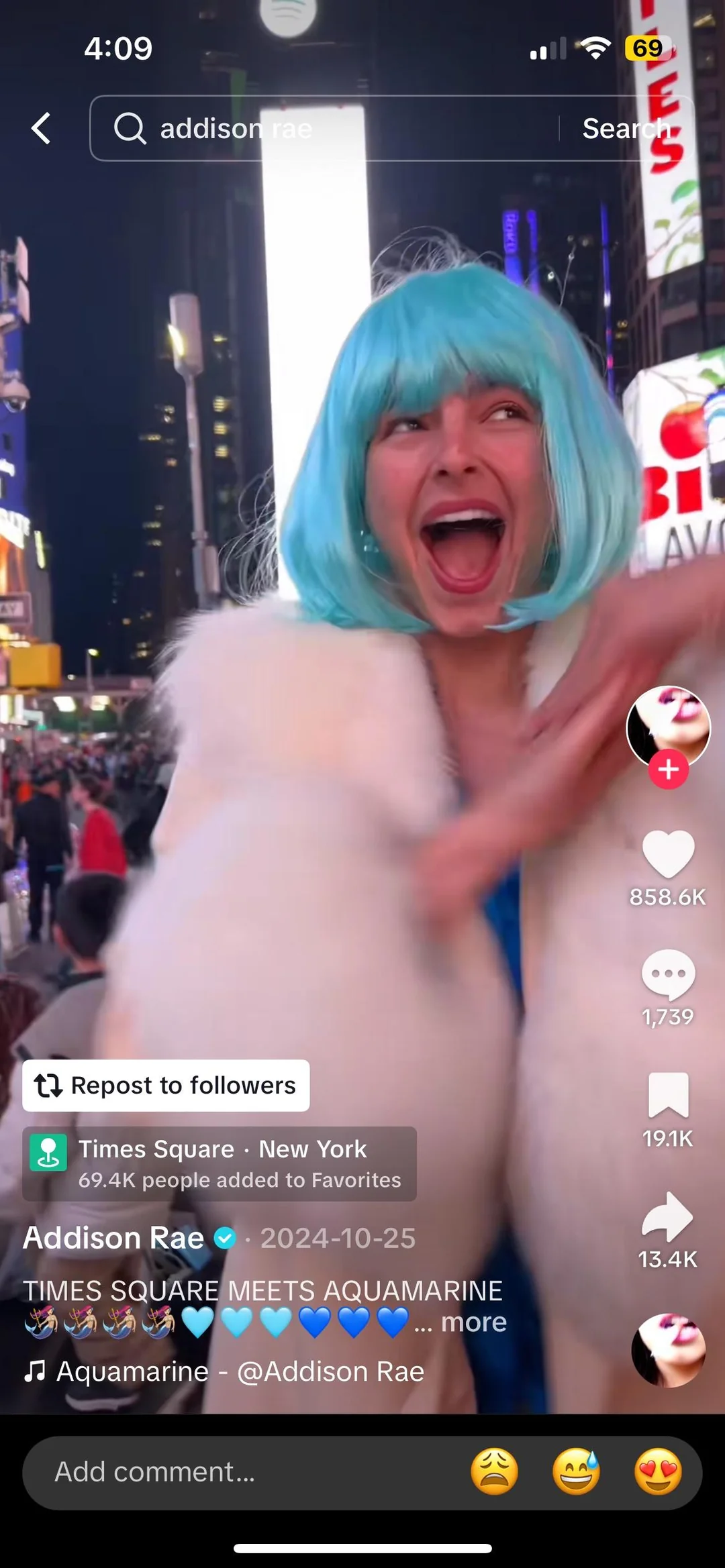 A smiling person with a bright blue wig and white fur coat poses excitedly in Times Square at night, surrounded by colorful lights and digital billboards. TikTok overlays and engagement icons are visible on the screen.