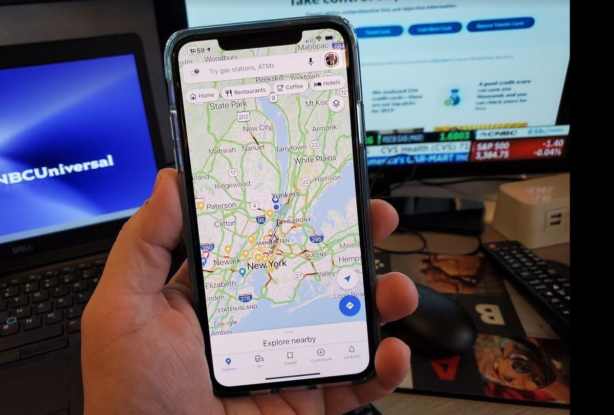A hand holds a smartphone displaying Google Maps with the New York City area shown; in the background, a computer screen and a TV with news channels are visible on a desk.
