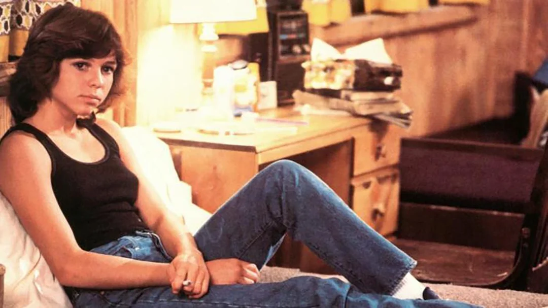 A person with short brown hair, wearing a black tank top and blue jeans, sits on a bed in a wood-paneled room with papers, a lamp, and books on a desk in the background.