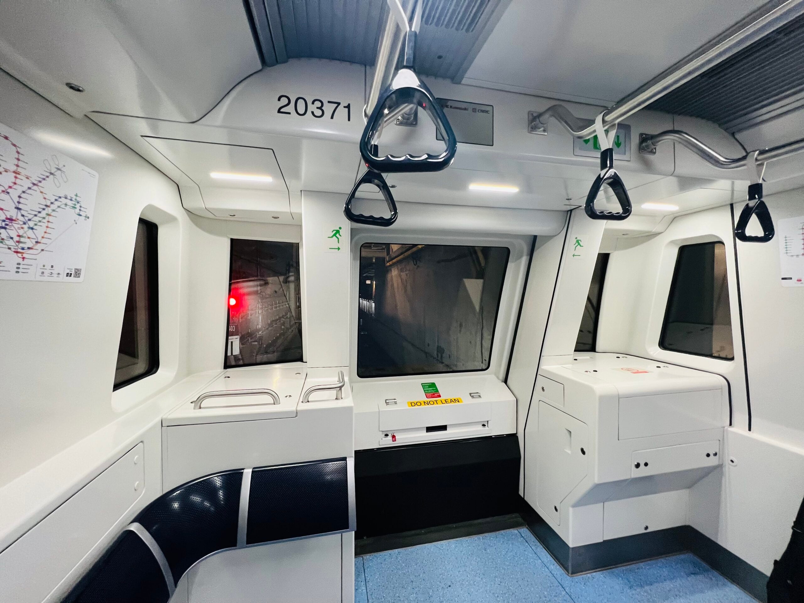 View from inside the front car of a modern, driverless subway train with large windows overlooking the tracks, a control panel, safety signs, and hand grips for standing passengers.
