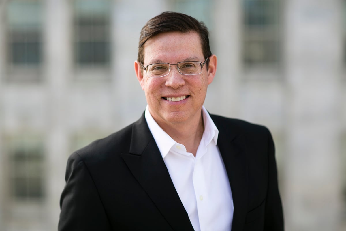 A man wearing glasses, a white shirt, and a black blazer is smiling outdoors in front of a blurred building background.