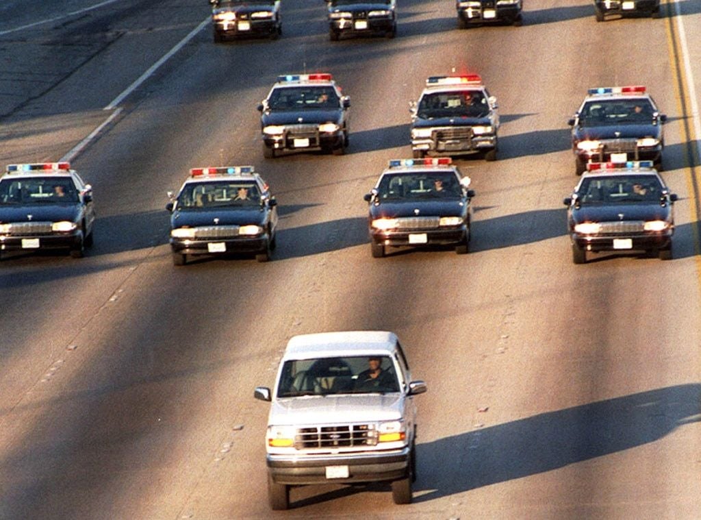 A white SUV is driving down a multi-lane highway, closely followed by several police cars with flashing lights. The scene appears tense, with police vehicles in pursuit formation behind the SUV.