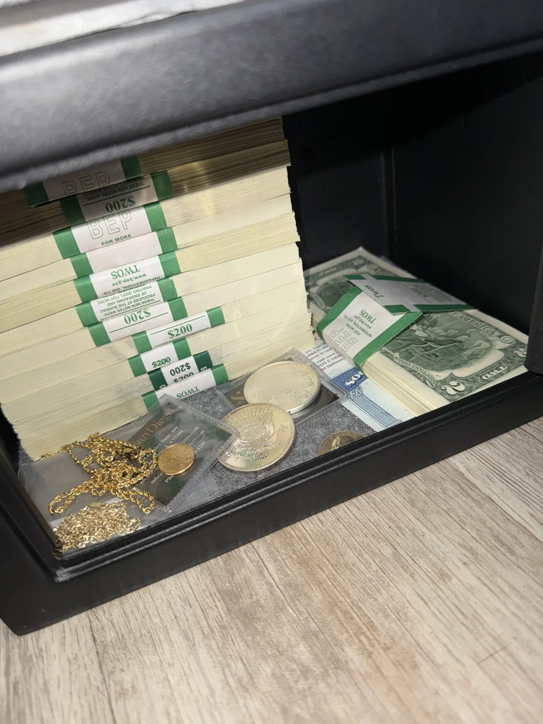 A safe containing stacks of U.S. $20 bills, gold jewelry, gold coins, silver coins, and other currency sits on a wooden surface.