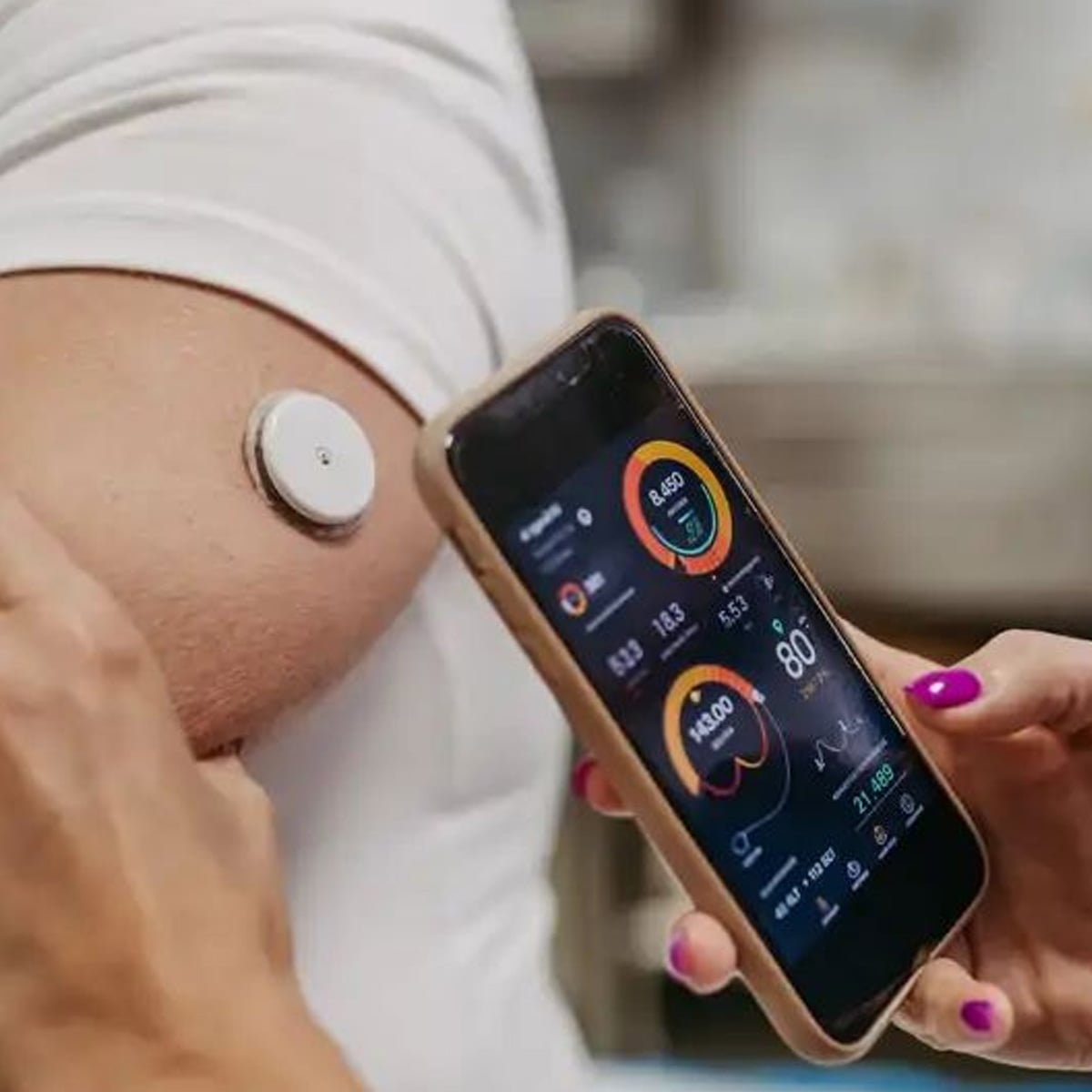 A person holds a smartphone displaying health data near their upper arm, where a round wearable sensor is attached, likely monitoring glucose or other health metrics.