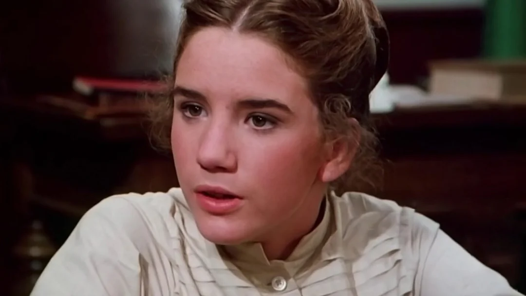 A young woman with brown hair styled up, wearing a high-collared, cream-colored blouse, sits indoors with a serious expression. Books and wooden furniture are visible in the blurred background.