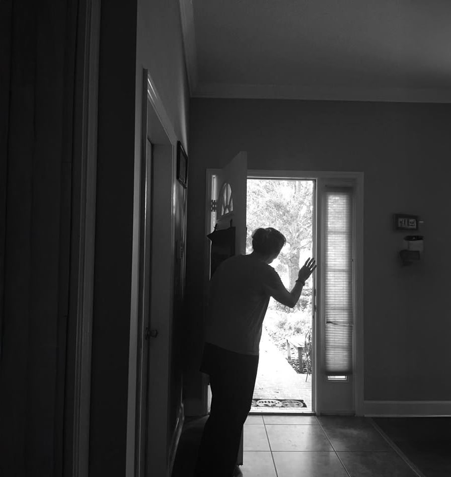 A person stands in the doorway of a house, leaning forward and waving outside. Sunlight streams in through the open door, creating a contrast with the dim interior. The scene appears calm and contemplative.