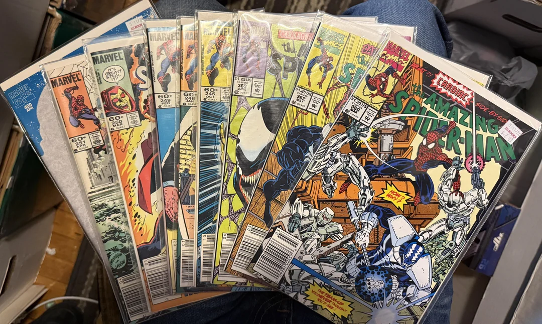 A person holds a fanned-out stack of vintage Marvel comic books, including several issues of The Amazing Spider-Man, all in protective plastic sleeves, with price stickers visible on the covers.