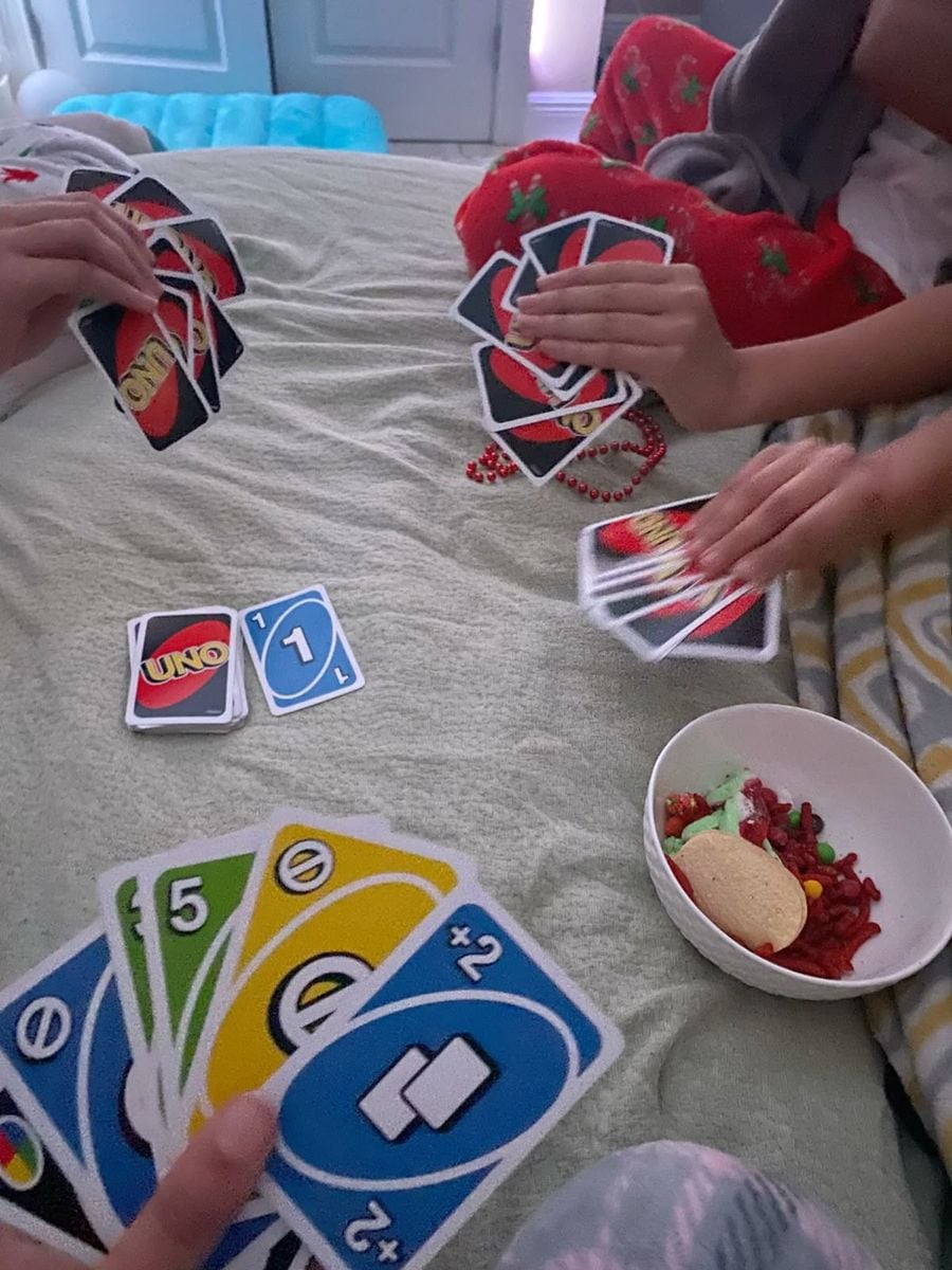Three people are playing an UNO card game on a bed with blankets; one holds green and blue cards. A small bowl of snacks, including cookies and candy, sits nearby. Pajamas and festive red clothing are visible.
