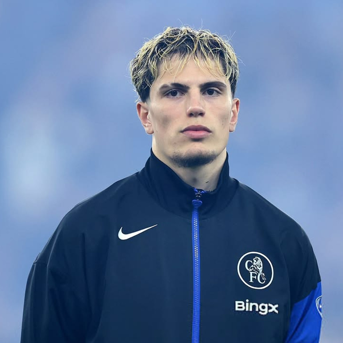 A young male footballer with short, blond-tipped hair wearing a black Chelsea FC jacket stands against a blurred blue background, looking directly at the camera with a neutral expression.