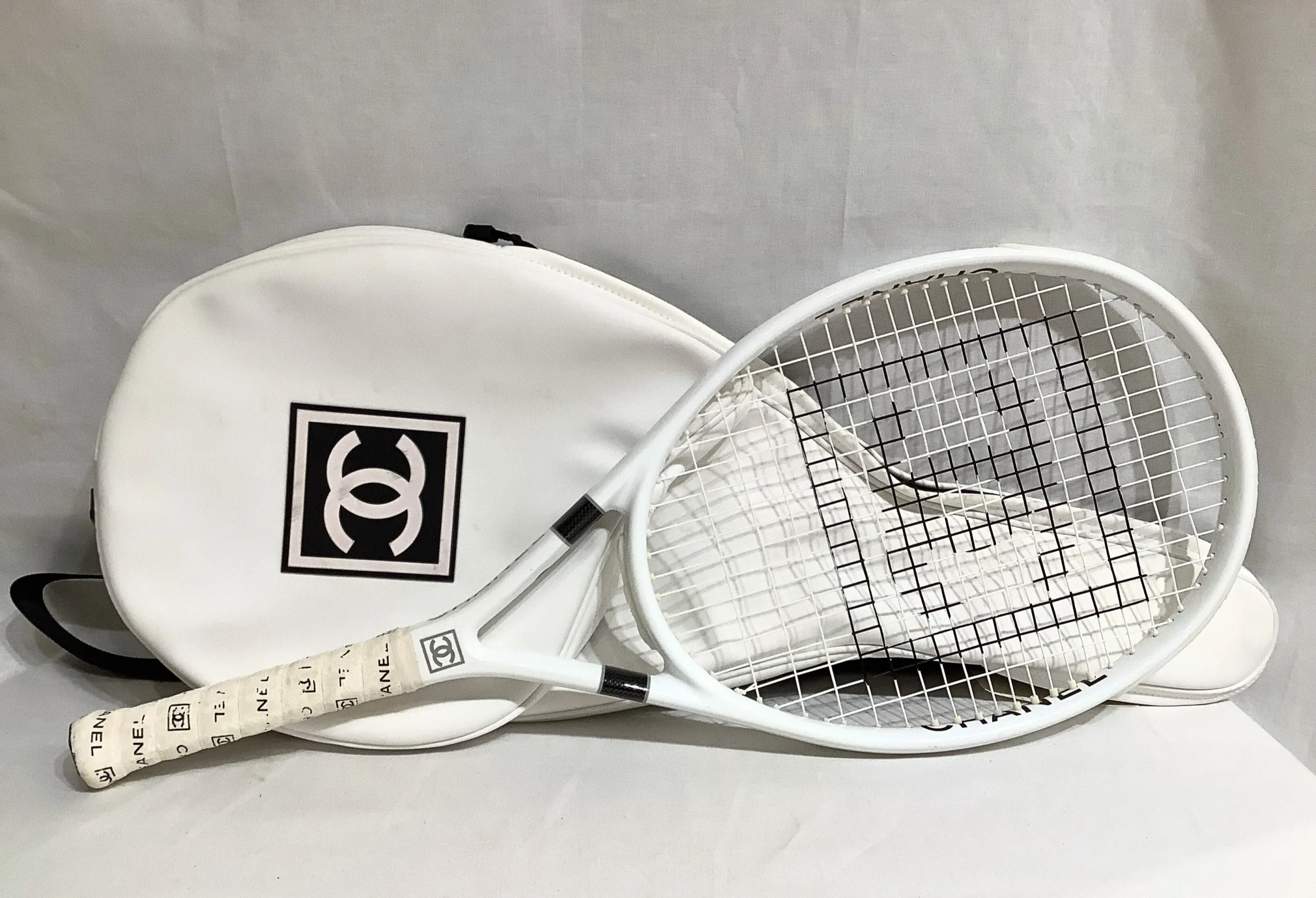 A white Chanel tennis racket with black accents rests on a matching white Chanel tennis bag, both featuring the iconic interlocking "CC" logo, against a neutral background.