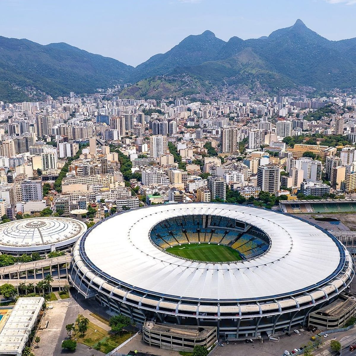 Aerial view of a large circular stadium surrounded by city buildings, with green mountains in the background under a clear sky. The stadium's field and seating are visible through its open roof.