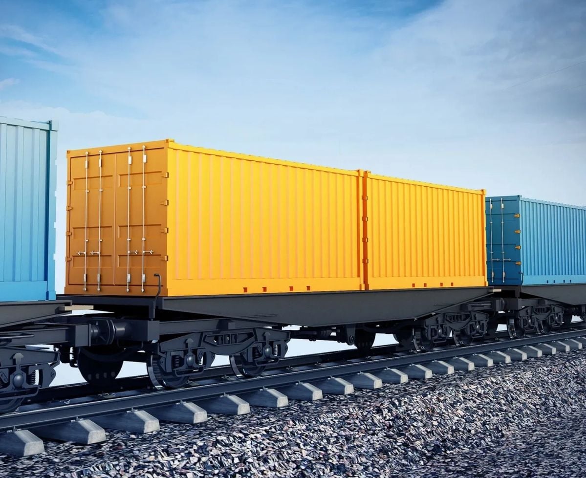 A row of colorful cargo containers, including yellow and blue, sits on flatbed train cars on railroad tracks under a blue sky.