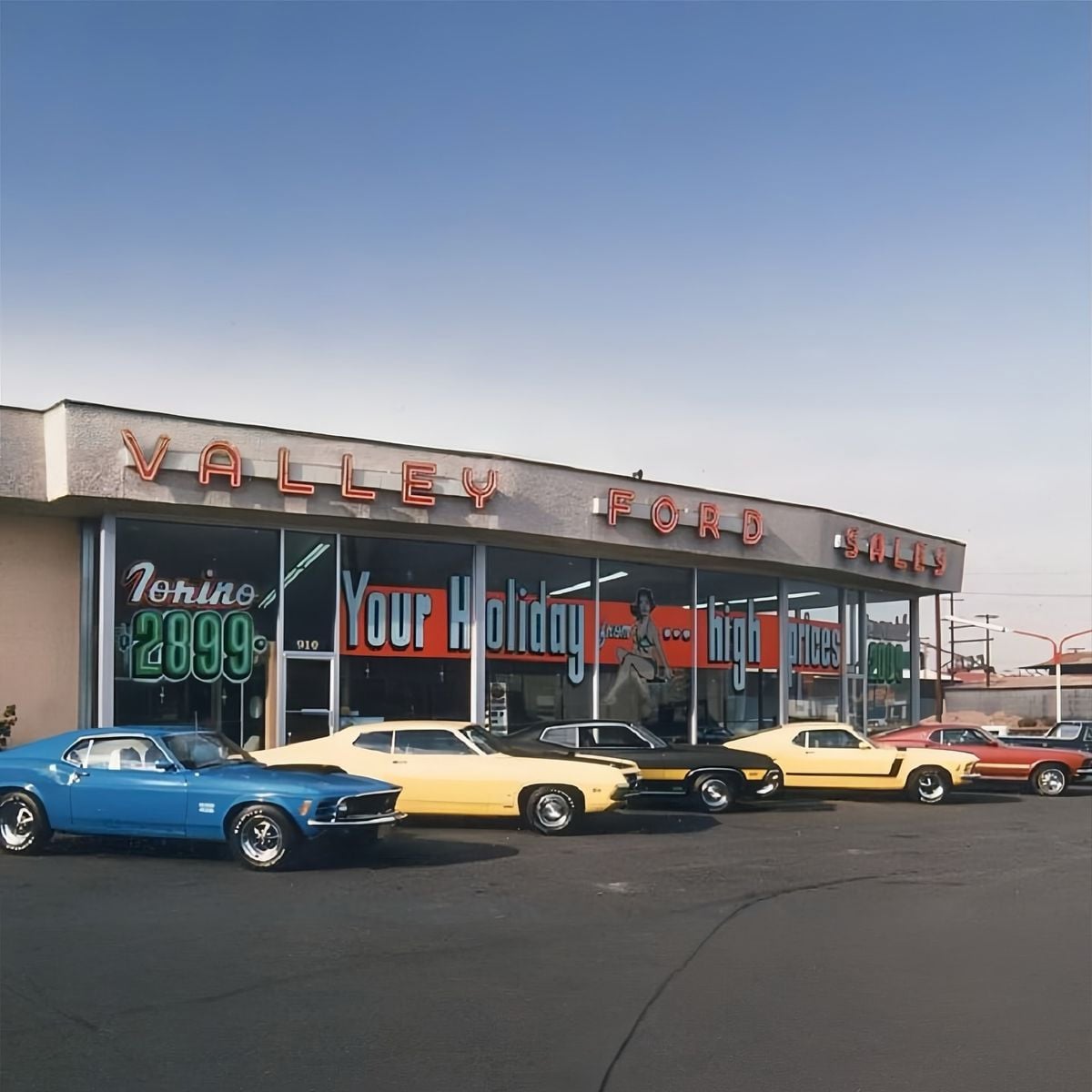 A row of classic 1960s cars in blue, yellow, black, gold, and red are parked in front of a Valley Ford dealership, with large glass windows and neon signage under a clear blue sky.