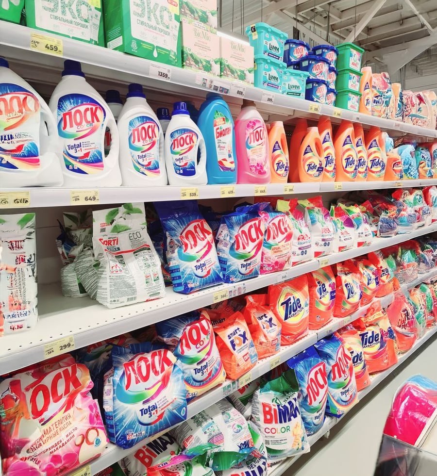 Shelves in a store stocked with various laundry detergents in bottles and bags, including brands like Losk, Tide, and Bimax, arranged in colorful packaging.