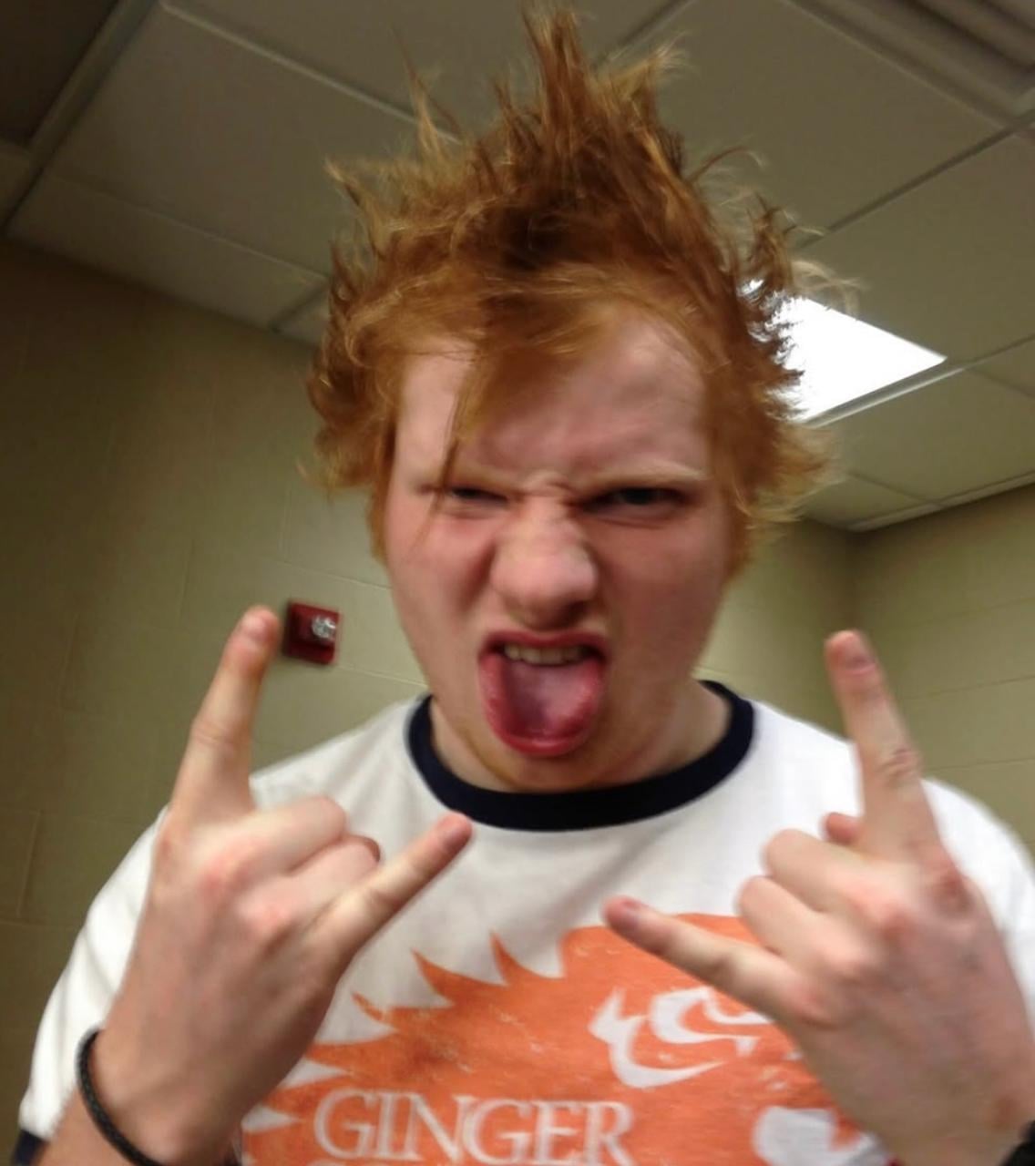 A person with spiky red hair makes a rock-and-roll hand gesture with both hands, sticks out their tongue, and scrunches their face, standing indoors under fluorescent lights.