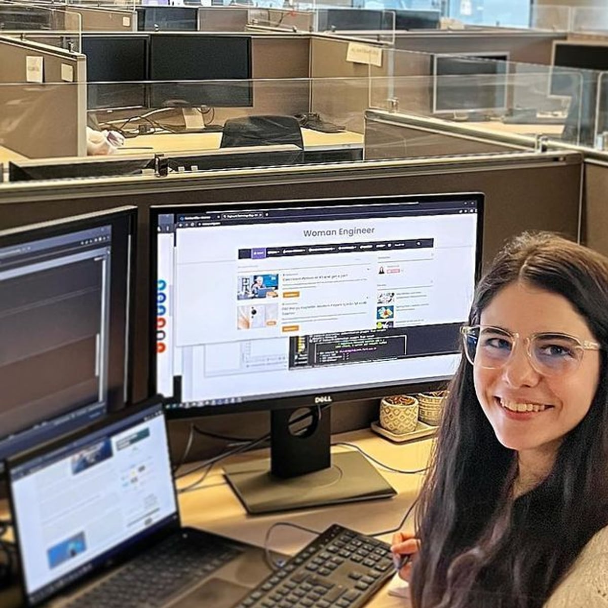 A woman with long dark hair and glasses smiles at a desk with a laptop and two monitors in an office cubicle. Her screen displays a webpage titled "Woman Engineer." Other empty cubicles are visible in the background.