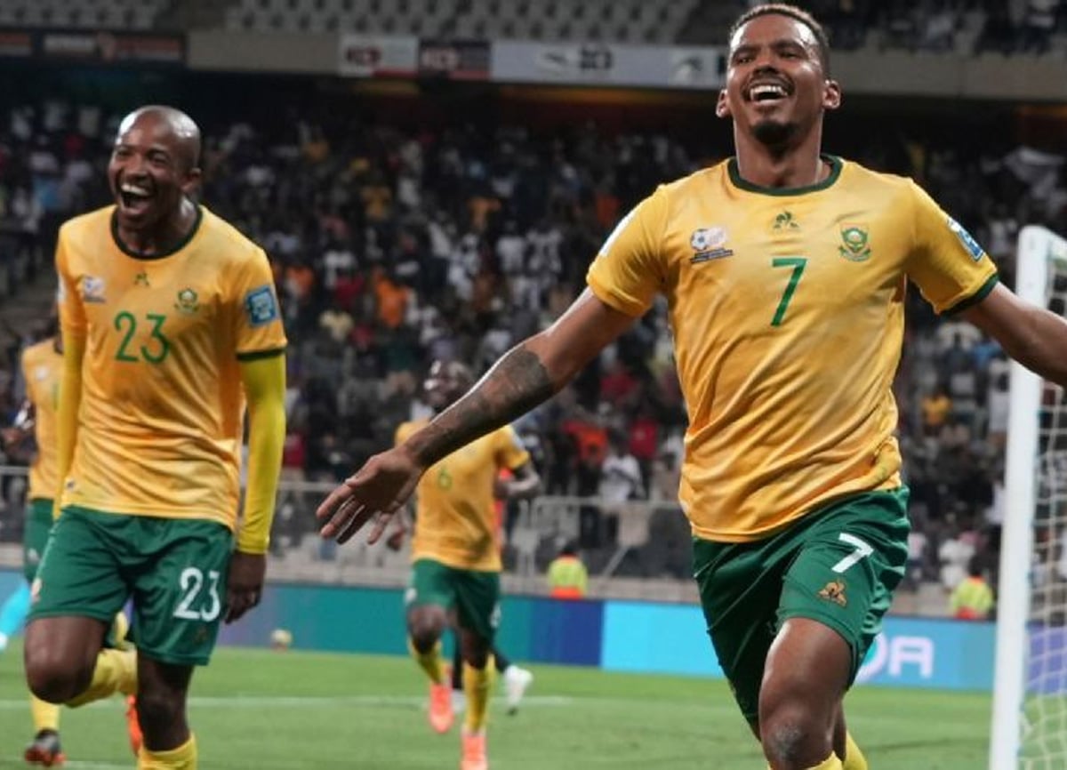 Two soccer players in yellow and green uniforms celebrate on the field, smiling with arms outstretched. The player in front wears number 7, while the player behind wears number 23. A crowd of spectators is visible in the background.