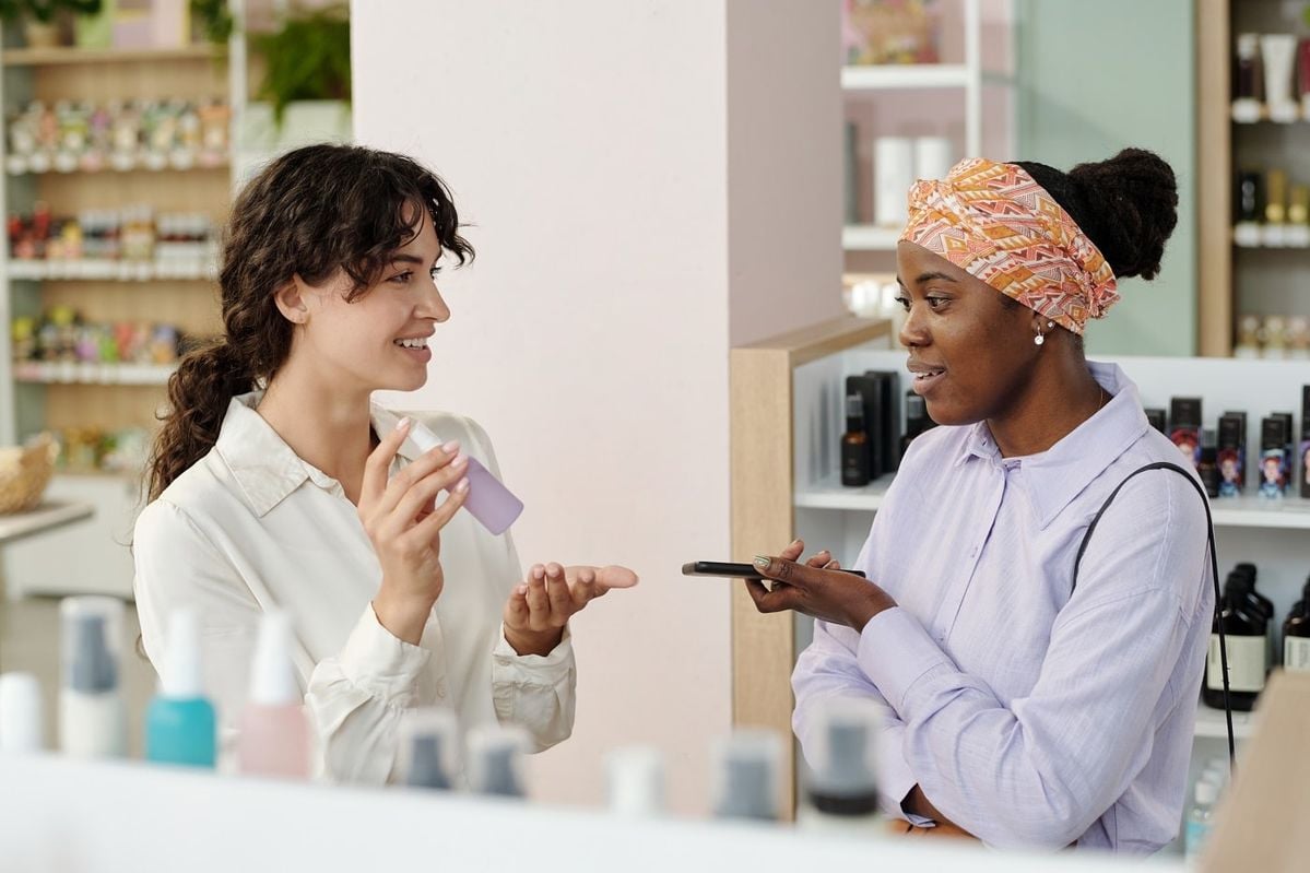 Two women in a store are talking; one holds a small bottle and gestures while smiling, and the other listens, holding her phone and wearing a headscarf. Shelves of products are visible in the background.