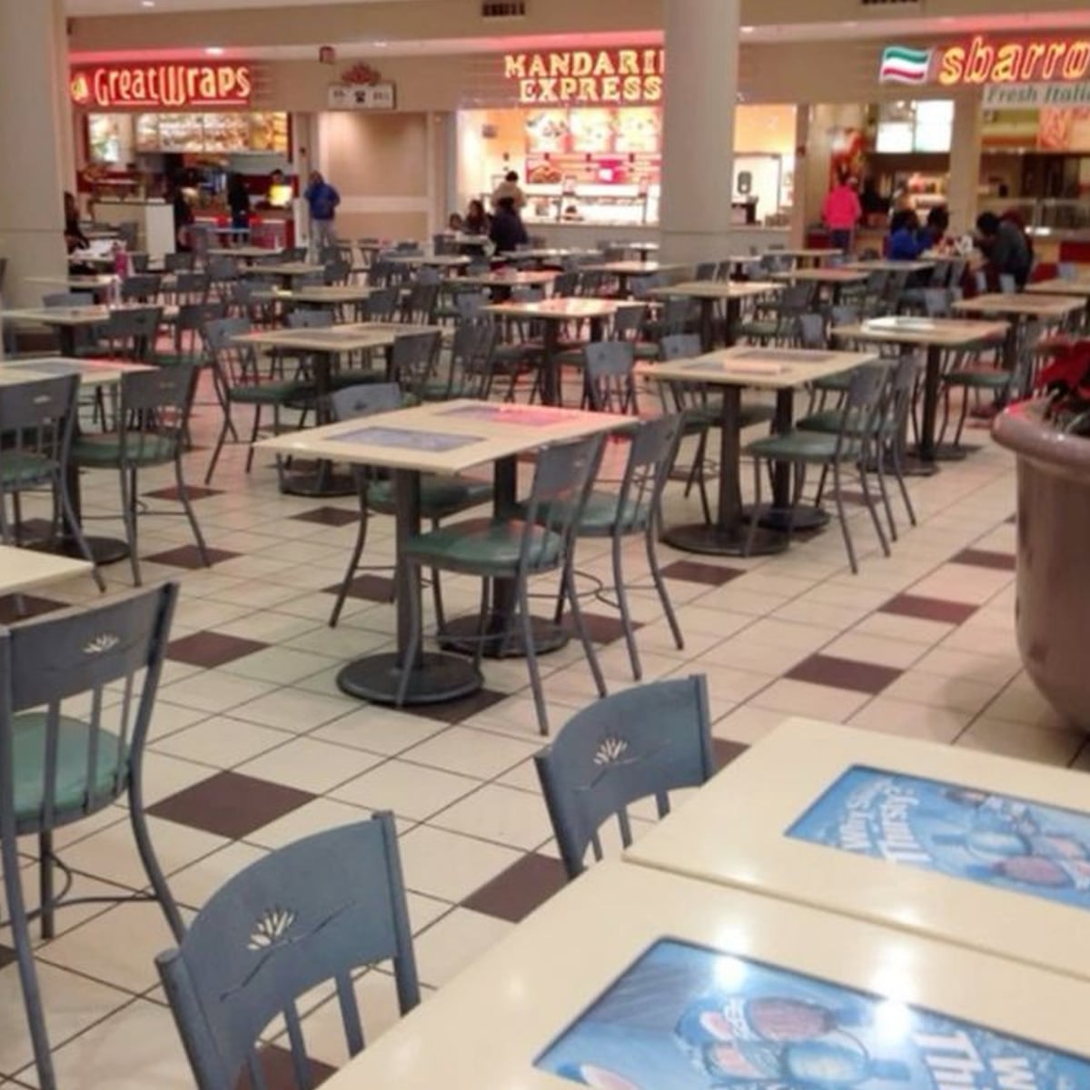 A mostly empty food court with rows of tables and chairs. Few people are seated in the background near restaurant counters like Great Wraps, Mandarin Express, and Sbarro. The floor is tiled.