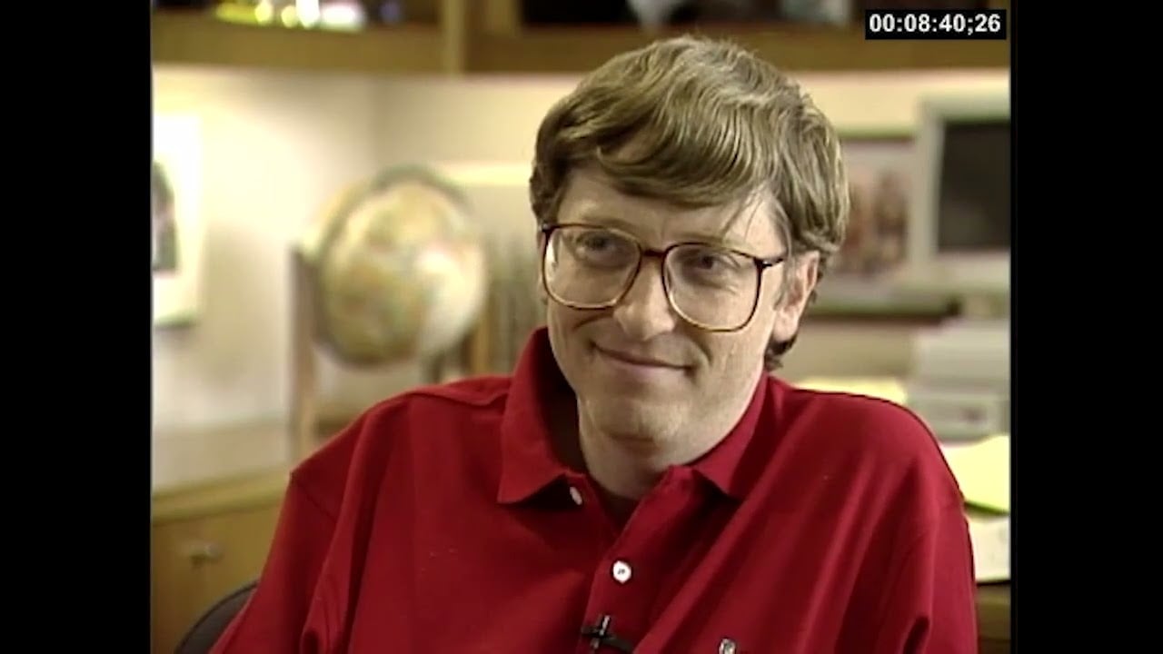 A man with glasses and light brown hair, wearing a red polo shirt, sits indoors and smiles slightly. There is a blurry globe and a computer in the background. A timestamp "00:08:40:26" appears in the top right corner.