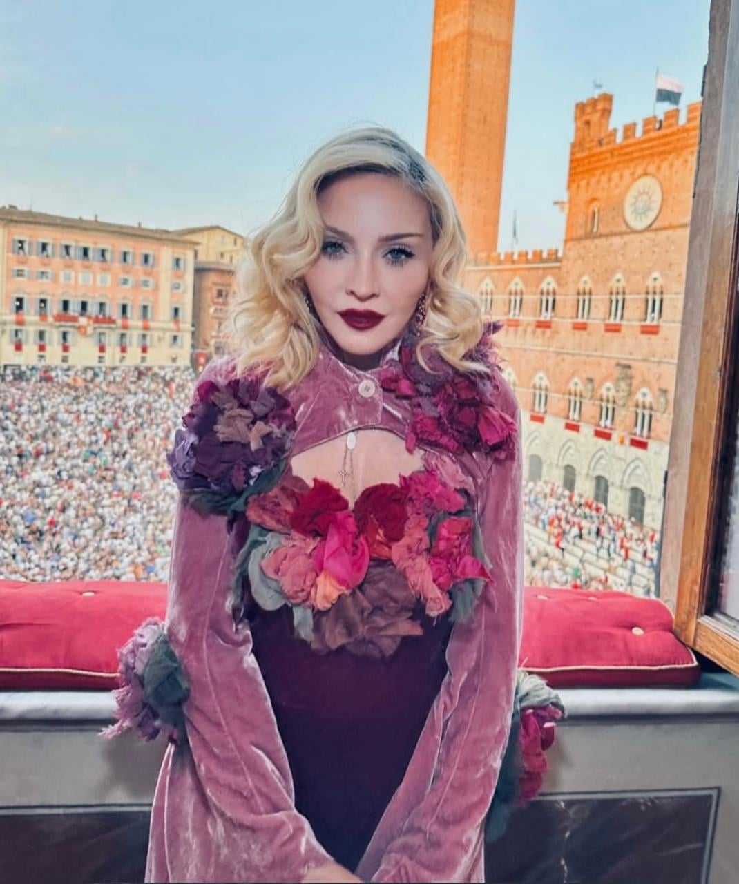 A woman with wavy blonde hair and deep red lipstick stands by a window, wearing a pink and purple floral-embellished outfit. A large crowd gathers in a historic square with old brick buildings in the background.