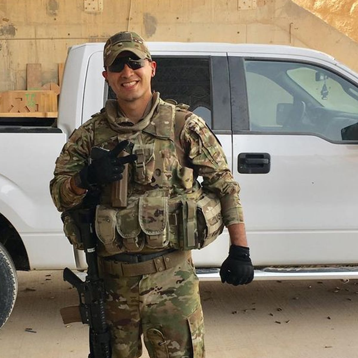 A smiling person in military camouflage stands in front of a white pickup truck, holding a beverage and flashing a peace sign. They are wearing sunglasses, gloves, and tactical gear, with a rifle slung across their body.