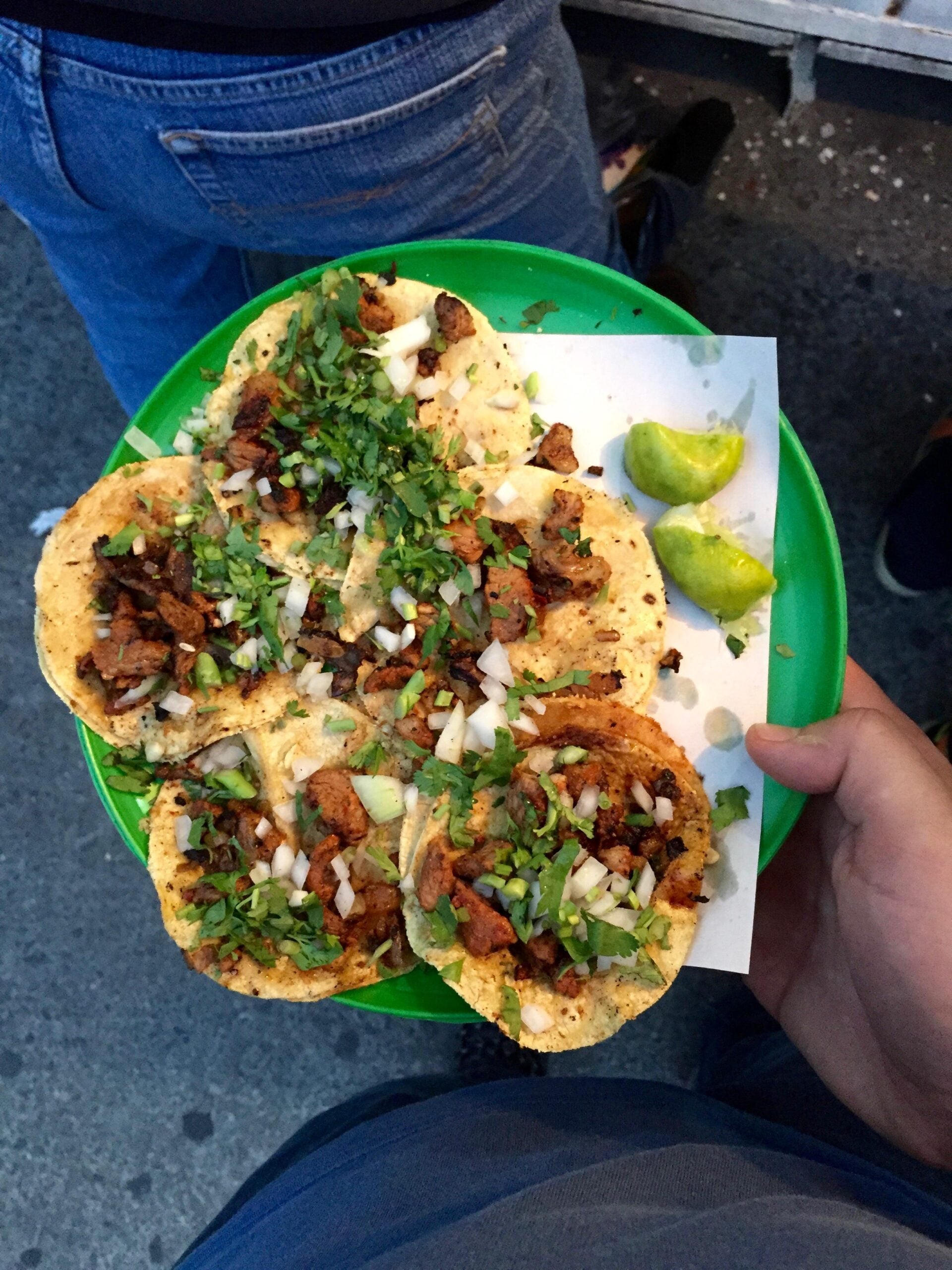 A person holds a green plate with five street tacos topped with chopped meat, onions, and cilantro, next to lime wedges, over a sidewalk background.