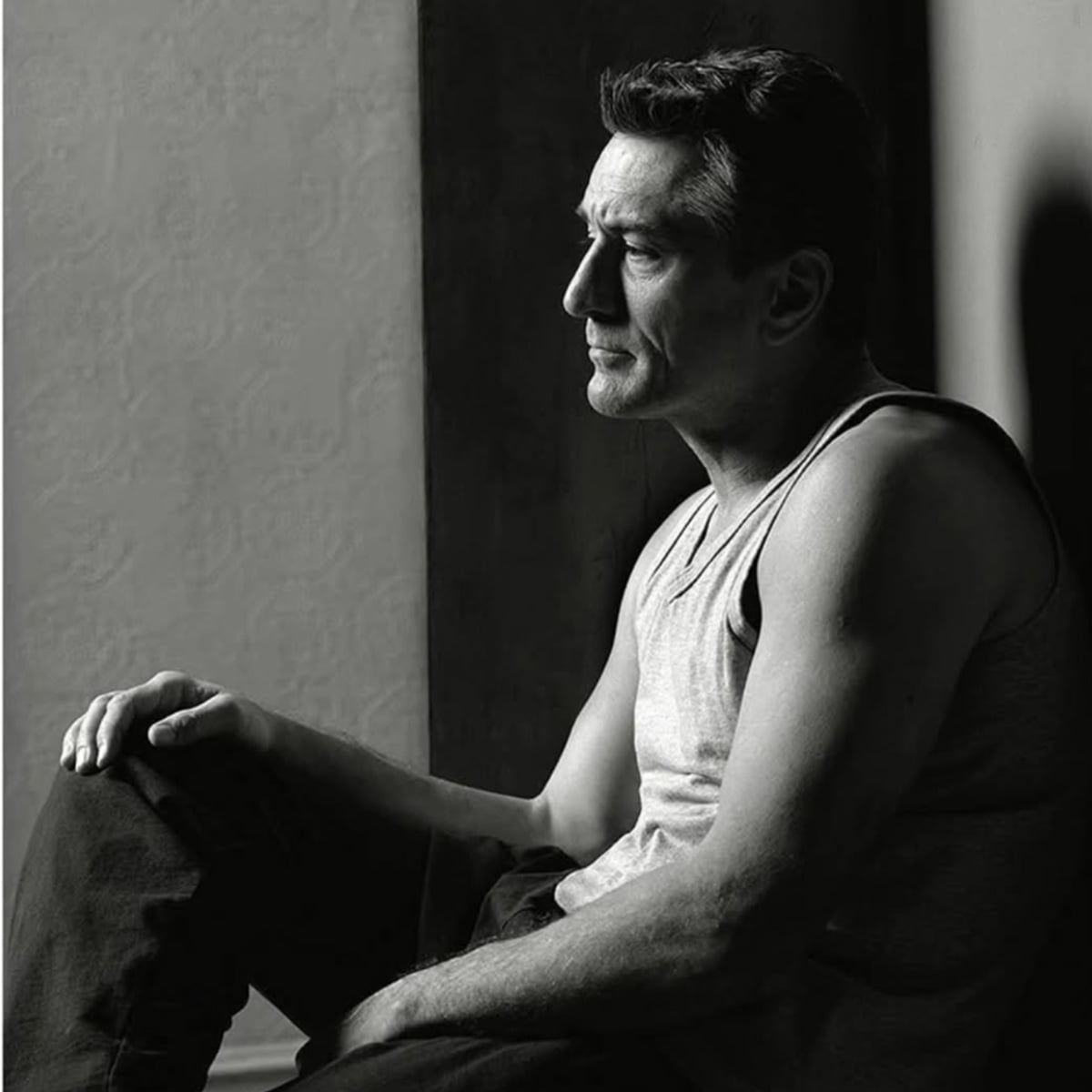 A man in a sleeveless shirt sits with one knee raised, gazing thoughtfully into the distance. The black-and-white photo highlights his contemplative expression and strong features, with soft light casting shadows on his face.