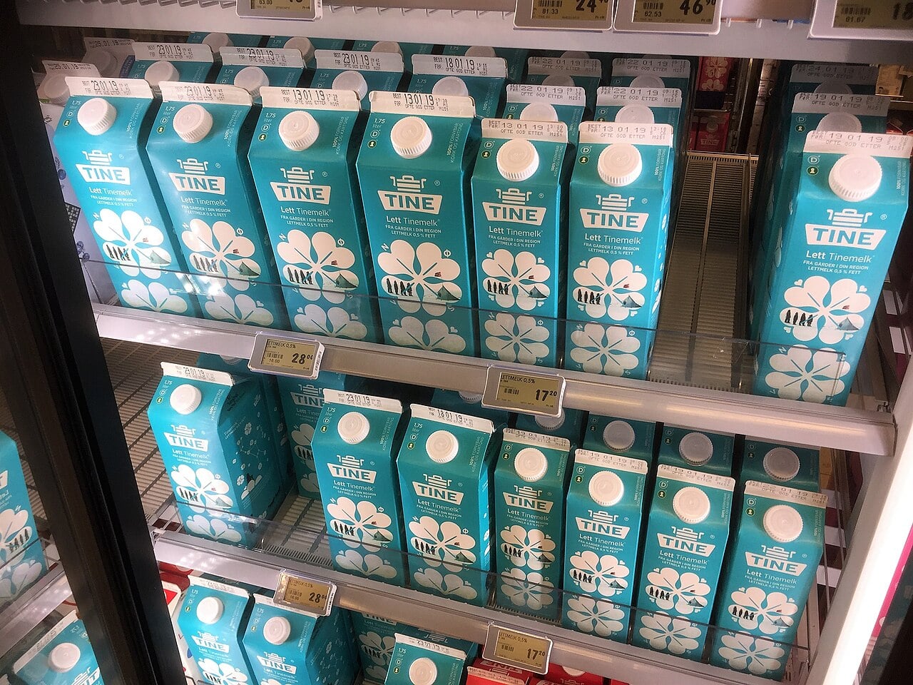 Blue and white milk cartons labeled "TINE Lett Tinemelk" are displayed on refrigerated shelves in a grocery store, with price tags visible on the shelf edges.