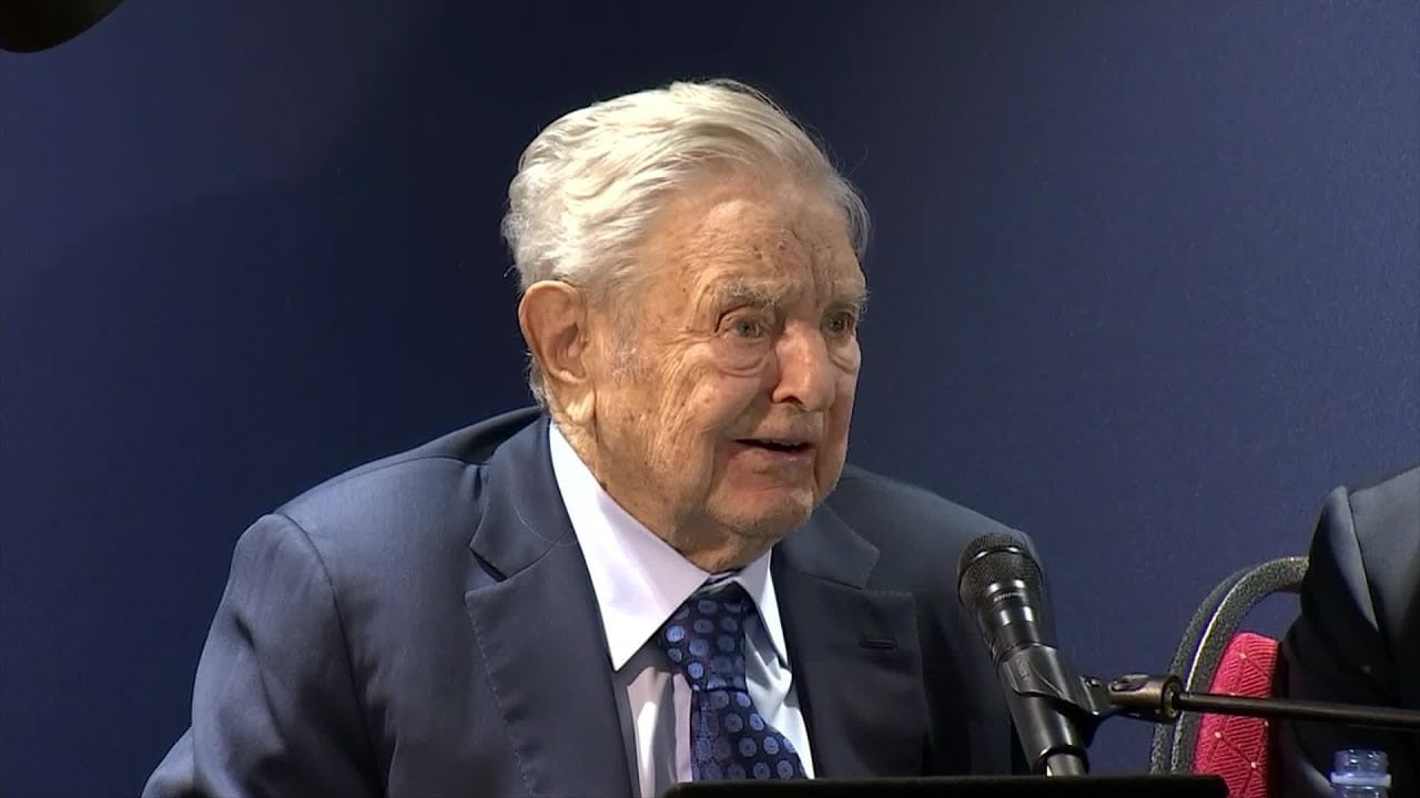 An elderly man in a suit and tie speaks into a microphone at a formal event, seated against a dark blue background.