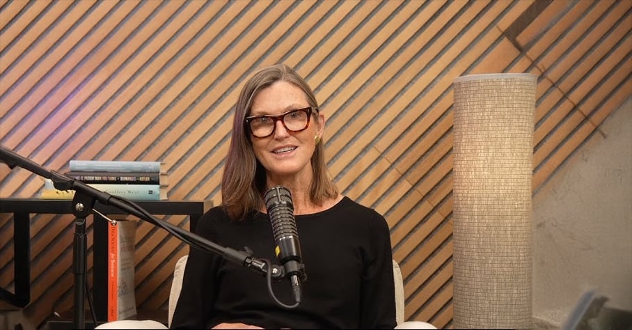 A woman with straight brown hair and glasses sits in front of a microphone, smiling. Behind her are stacked books on a black table, a tall textured lamp, and a wood-paneled wall with diagonal lines.