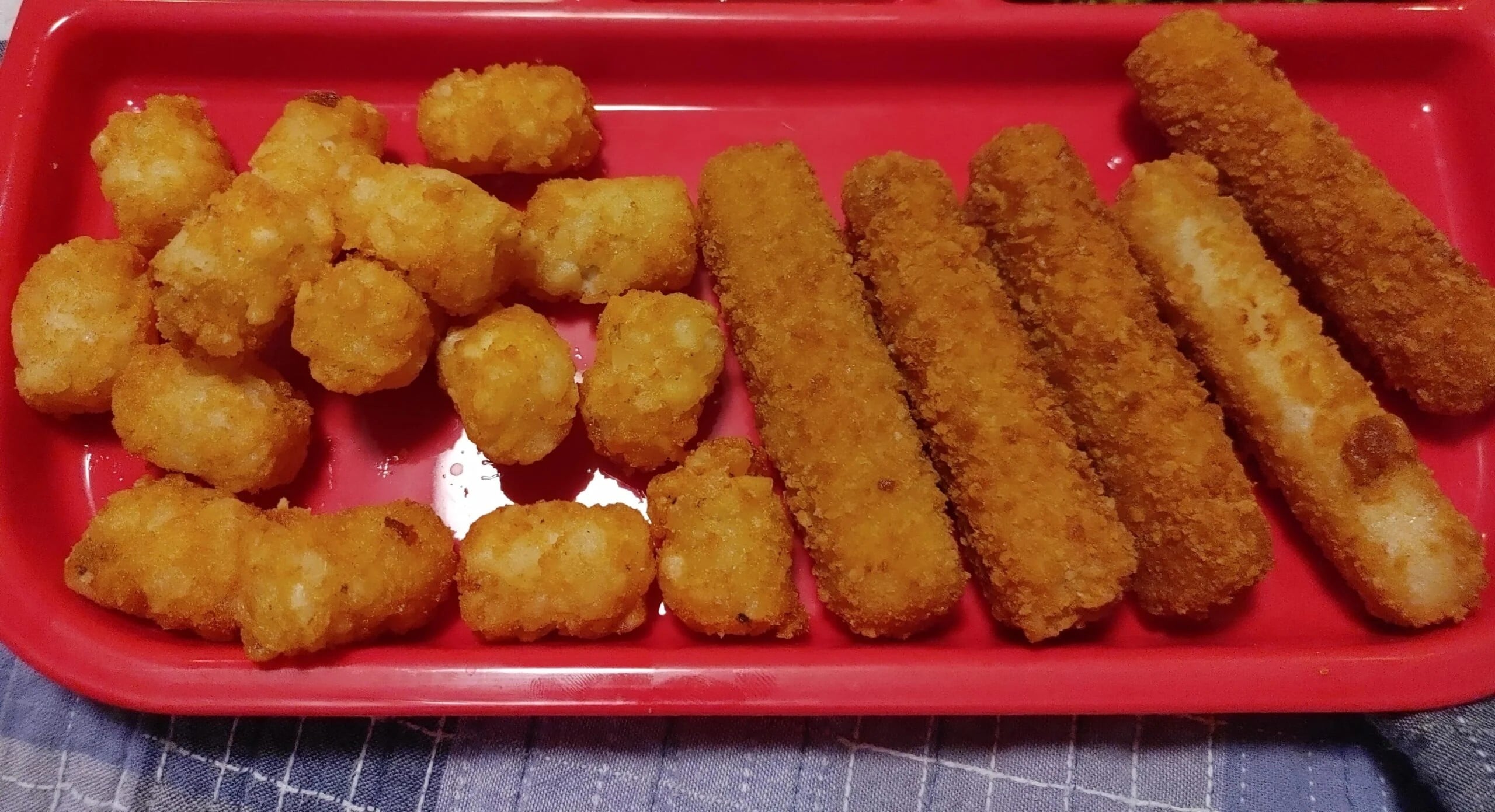 A red tray with a serving of golden-brown tater tots on the left and several breaded fish sticks lined up on the right, all placed on a blue and white patterned cloth.