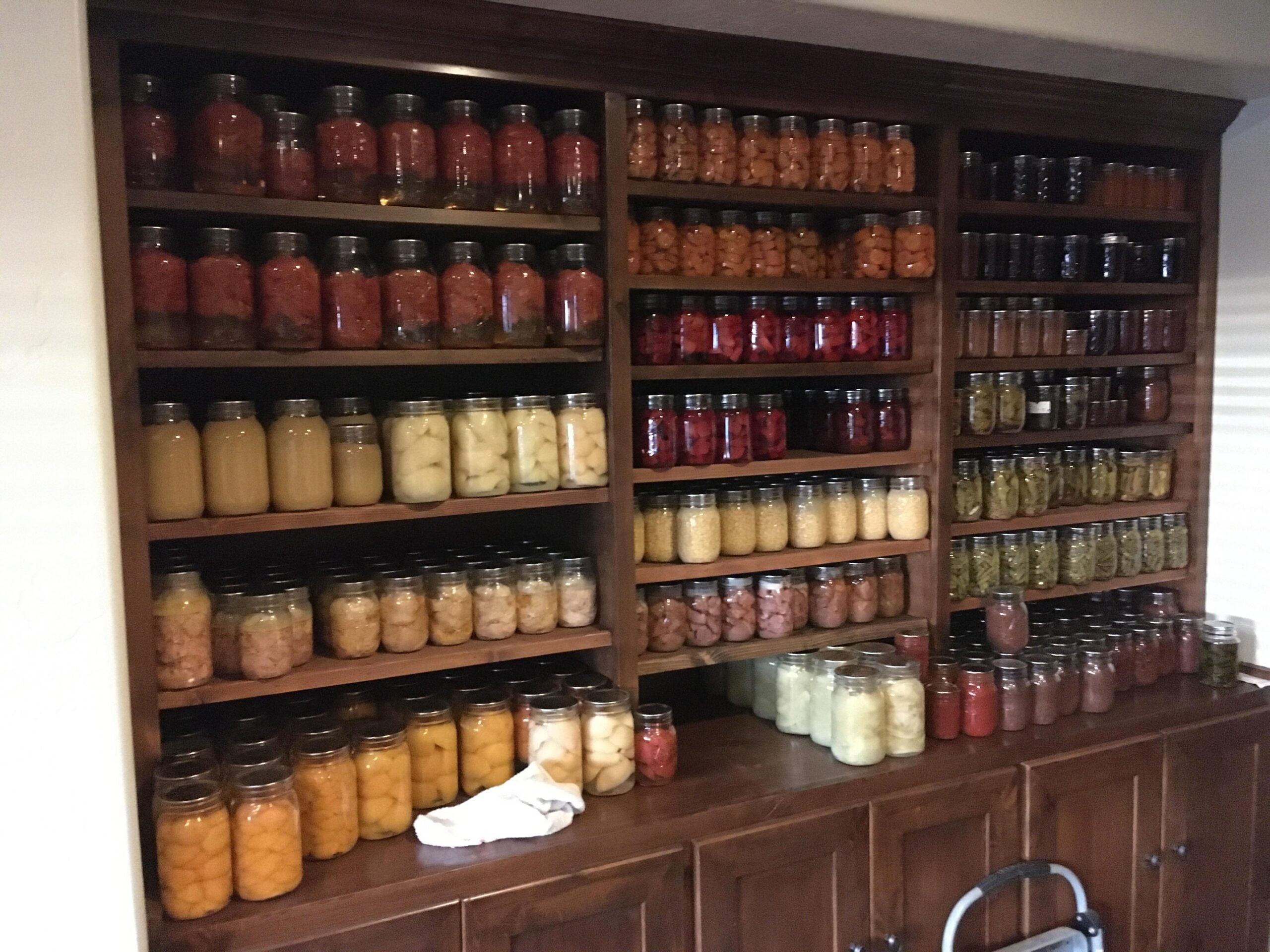 Wooden shelves filled with neatly organized jars of home-canned fruits and vegetables of various colors, including peaches, pears, beans, tomatoes, and other preserved foods. A few jars and a towel sit on the counter below.