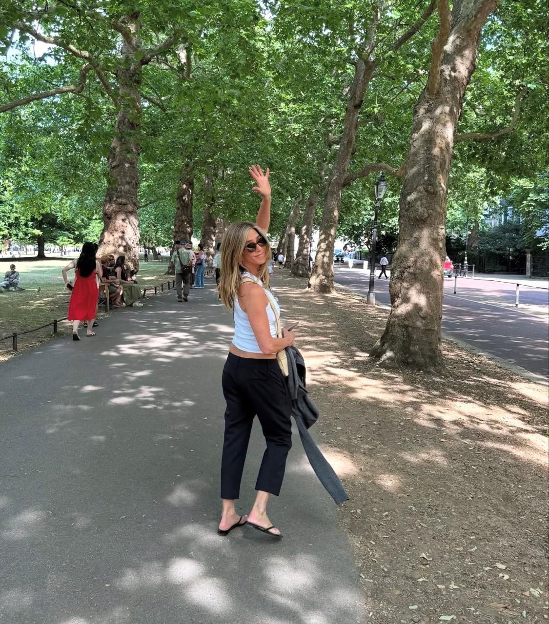 A woman wearing sunglasses, a white top, and black pants stands on a tree-lined path, looking back and waving. Other people walk or sit nearby in a green, sunny park.