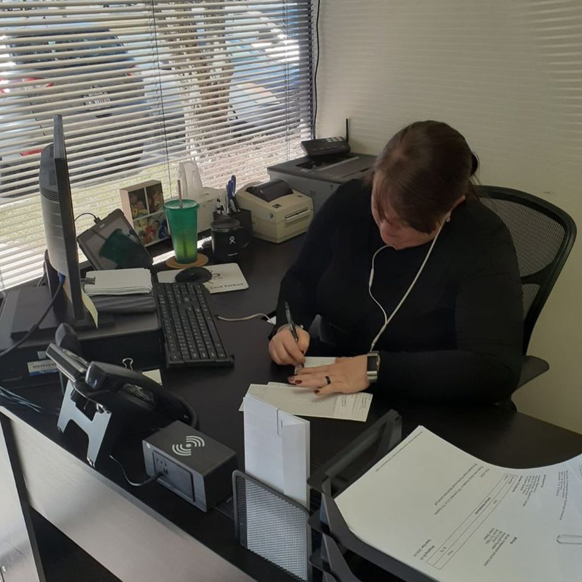 A person with brown hair tied back is sitting at a desk, writing on paper. The desk has a computer monitor, keyboard, phone, and various office supplies. Sunlight comes through a window behind them.