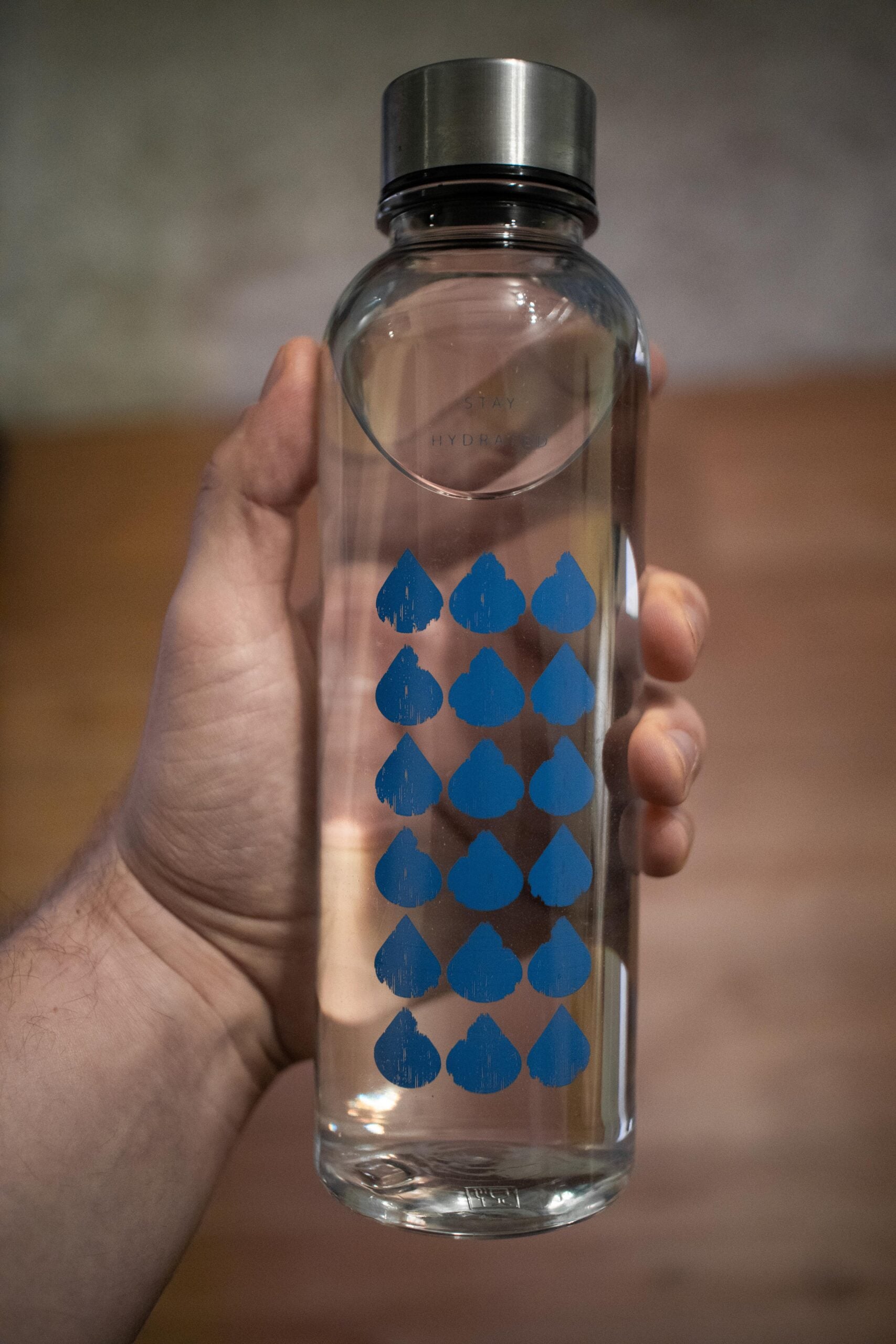 A hand is holding a clear glass water bottle with a metal cap. The bottle has rows of blue water droplet icons printed on it. The background is blurred, showing brown and gray tones.