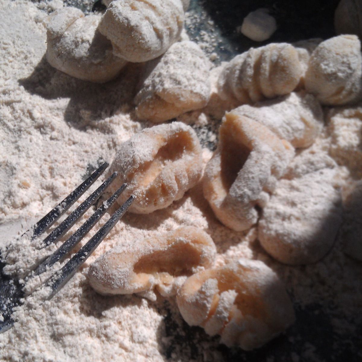 Uncooked gnocchi pieces dusted with flour are scattered on a surface, with a fork used to shape their ridges resting nearby. Bright light highlights the texture of the dough and flour.