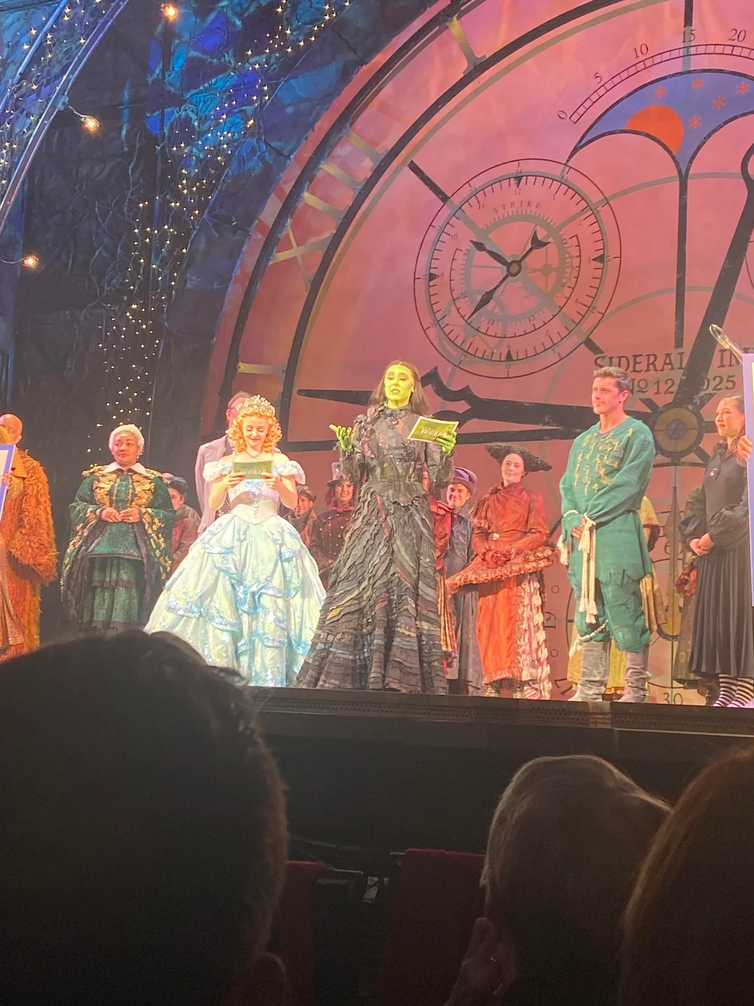 Actors in colorful, elaborate costumes take a bow on stage after a performance. The central character is dressed in black with green face paint, while another wears a blue gown. A large clock set is in the background.